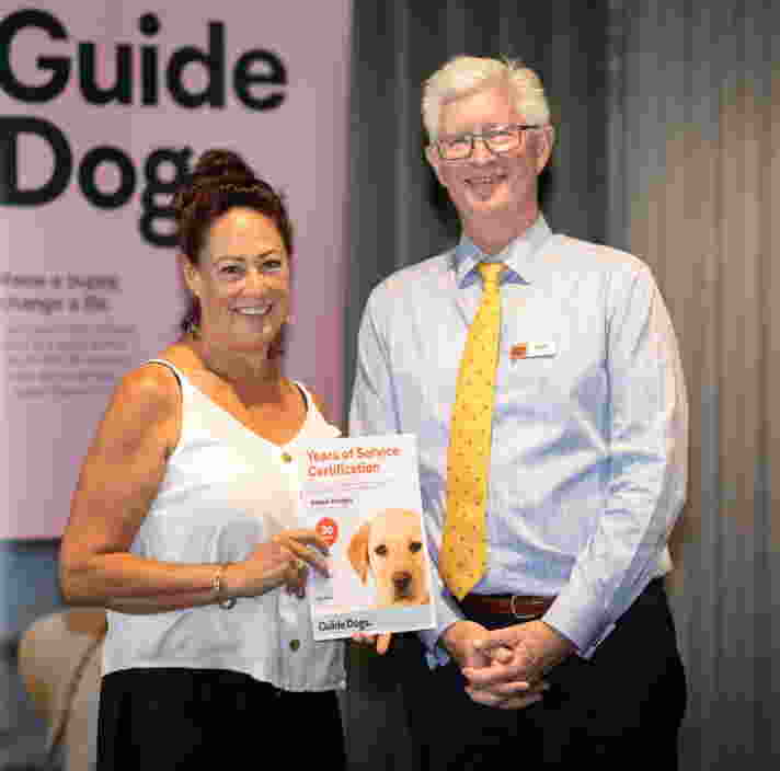 Two people standing in front of a pink banner with the Guide Dogs logo. The people are both smiling at the camera and one is holding a certificate.