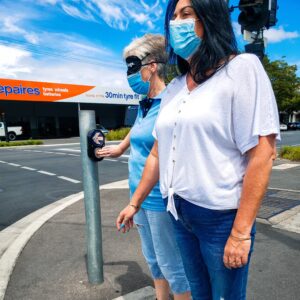 Two women standing at a traffic light. One woman is blindfolded and pressing the traffic light button.