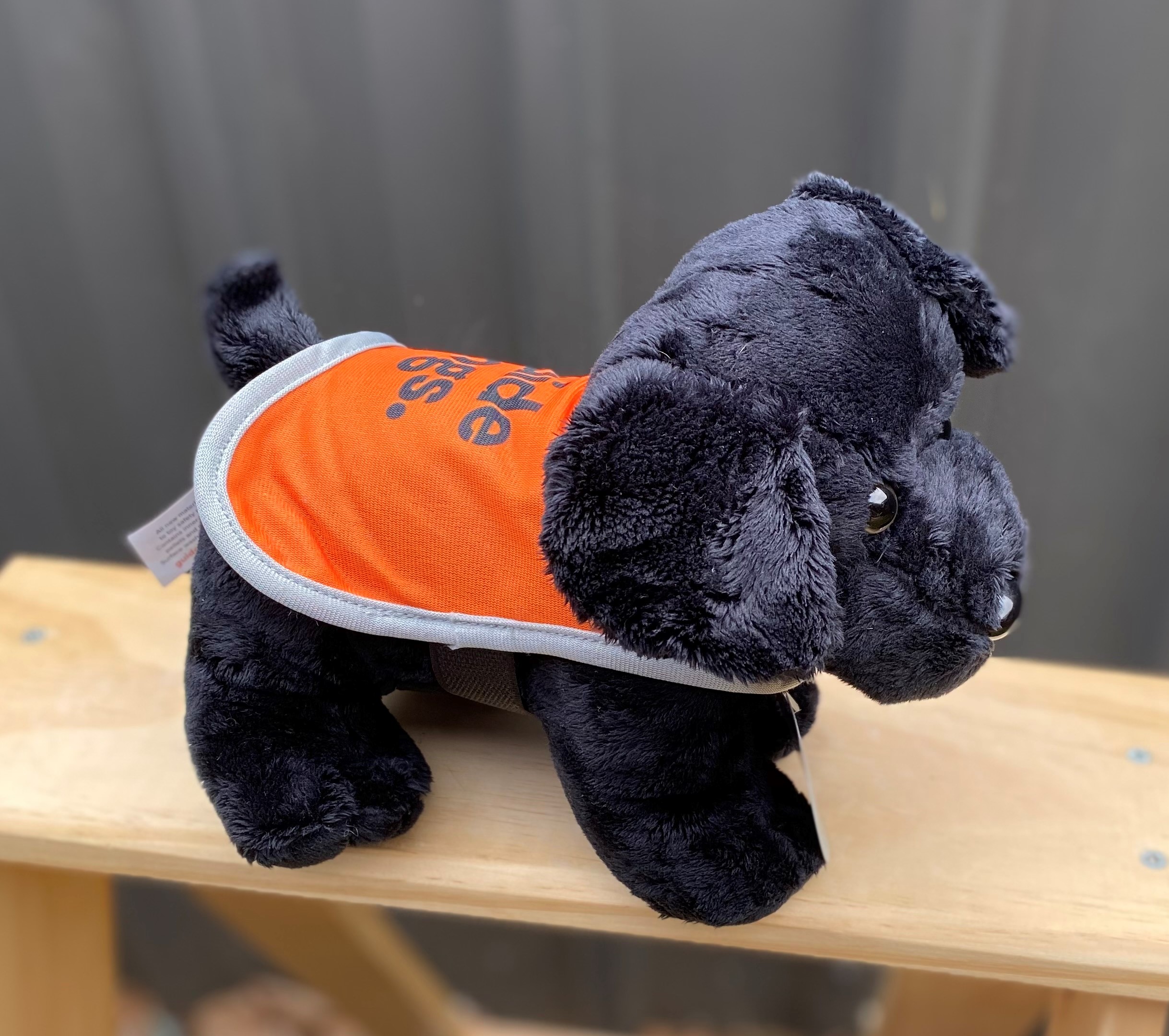 The Plush Dog - Standing, a black plush dog toy in an orange vest, is positioned on a wooden surface.