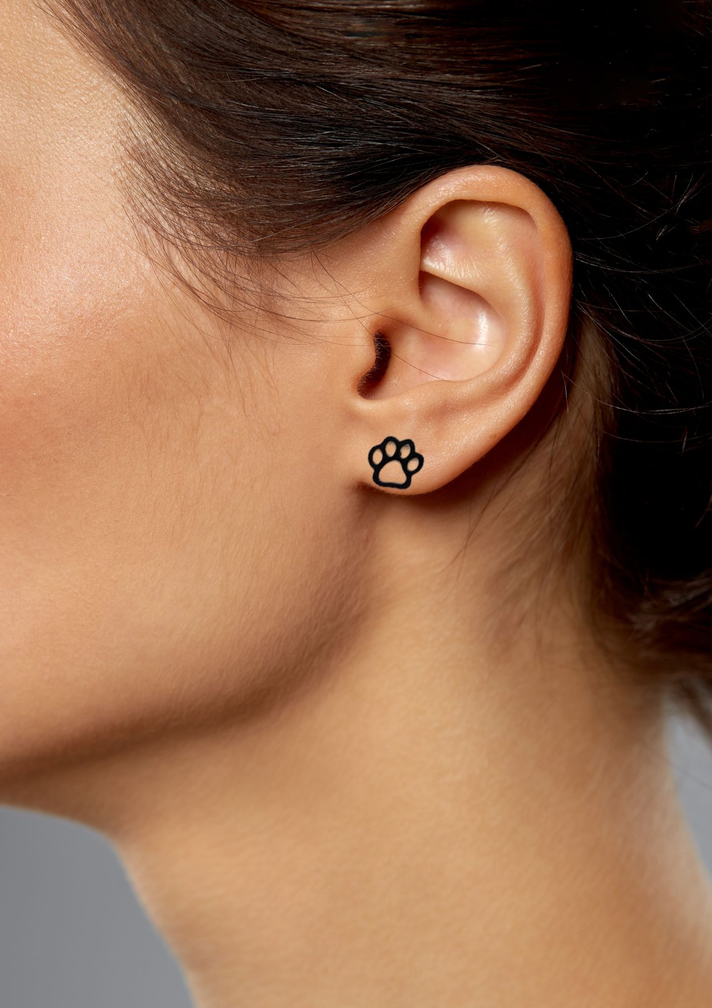A close-up shot of someone's ear adorned with a small black earring in the shape of a paw print, set against a gray background.