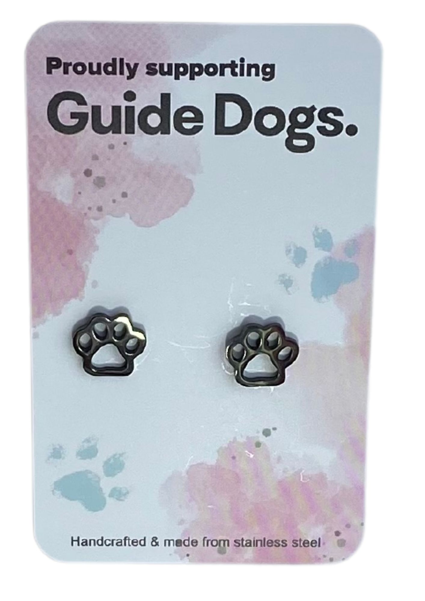 The image shows a silver paw-shaped earring on a card that reads "Proudly supporting Guide Dogs." The card has a pastel background with pink and blue splotches and small paw print designs. At the bottom, it states, "Handcrafted & made from stainless steel."