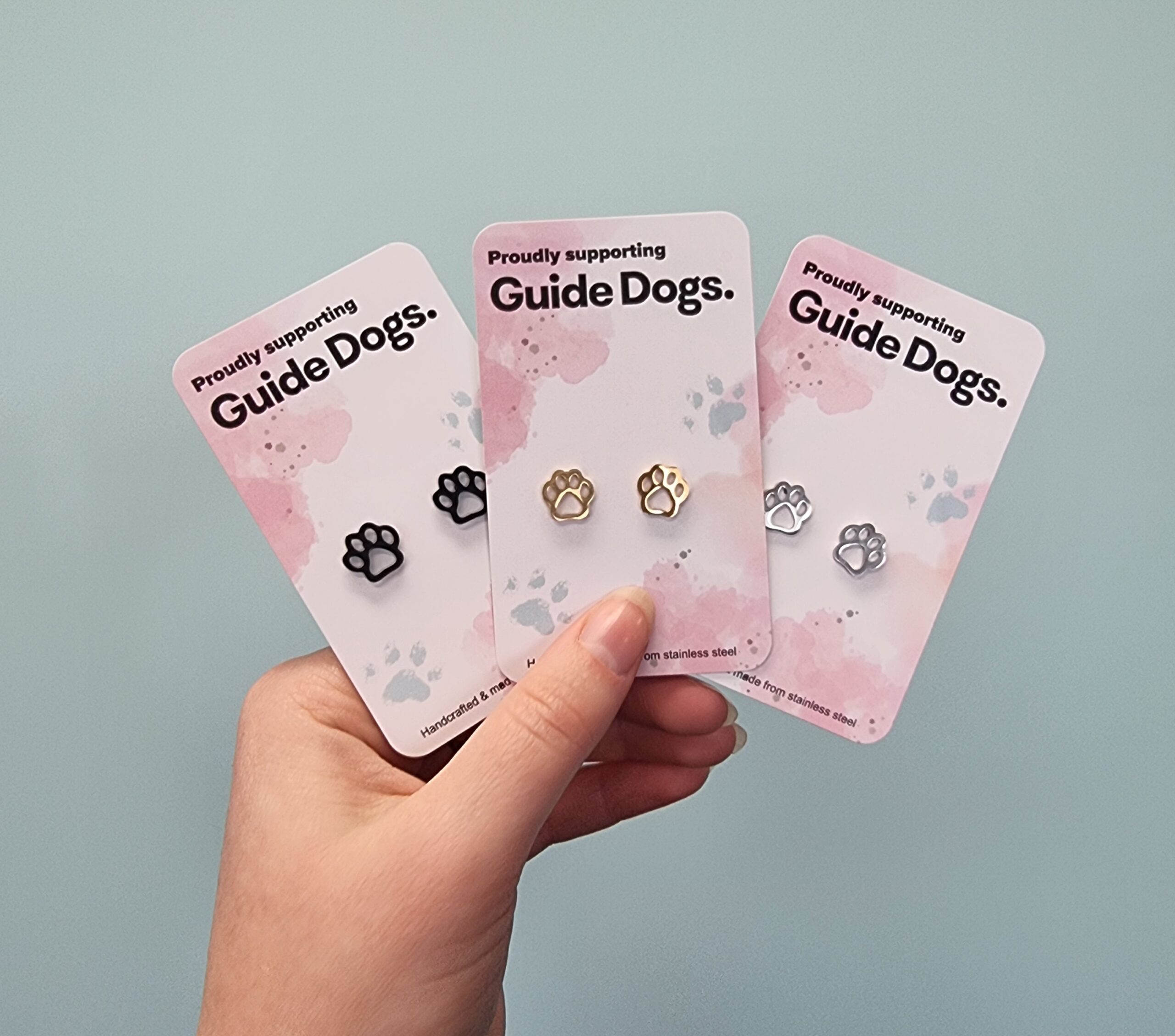 Image shows a hand holding three pairs of paw-shaped stud earrings, each displayed on a card that says "Proudly supporting Guide Dogs." The left pair is black coloured, the middle pair is gold and the pair on the right is silver coloured. The background is a plain light blue wall.