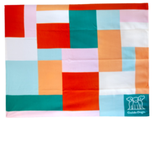 The Polar Fleece Rug is a colorful patchwork blanket adorned with pink, orange, teal, and white squares and rectangles. It features a small Guide Dogs logo in the bottom right corner.