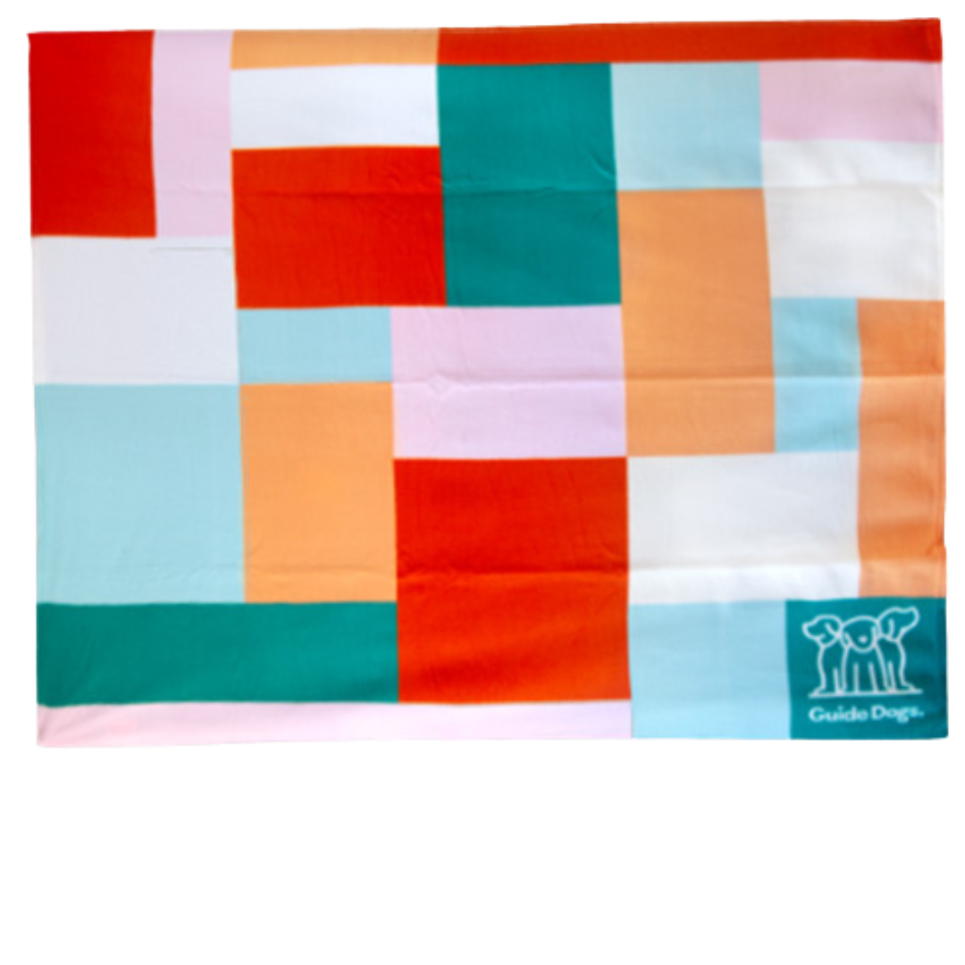 The Polar Fleece Rug is a colorful patchwork blanket adorned with pink, orange, teal, and white squares and rectangles. It features a small Guide Dogs logo in the bottom right corner.
