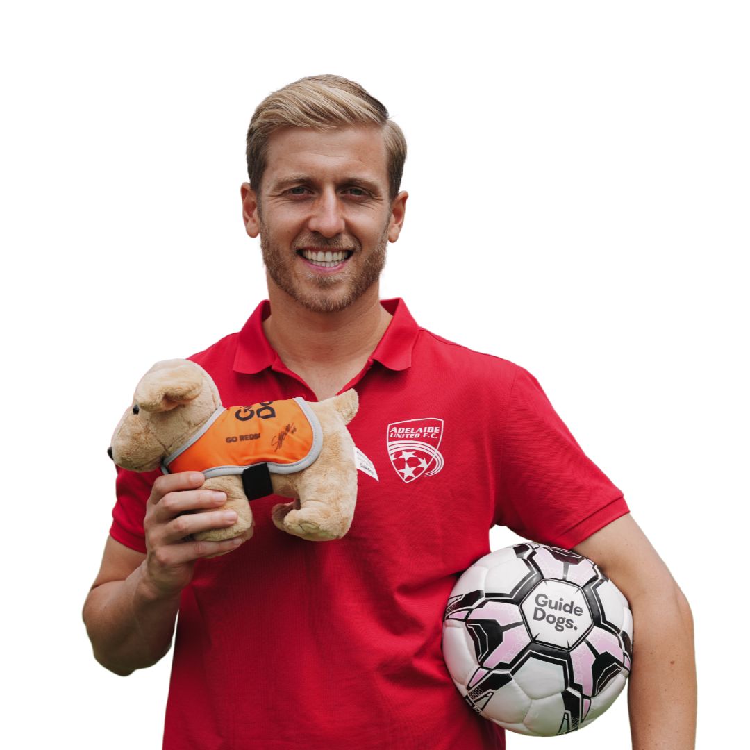 Man in a red shirt holding a cream plush dog and a soccer ball with Guide Dogs text.