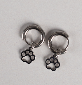 The Earrings - Huggie Paws feature silver hoops with black paw print charms, shown on a light gray surface.