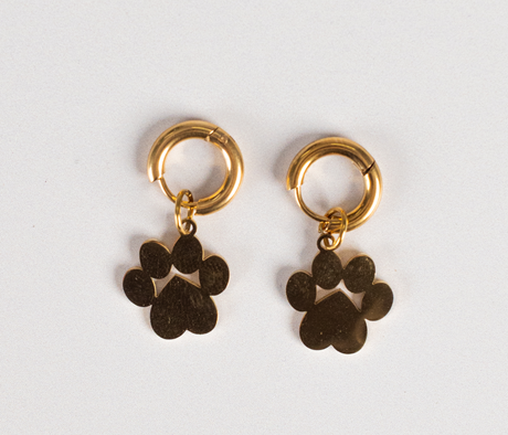 The Earrings - Huggie Paws, two gold hoops each with a dangling paw print charm, are displayed on a plain white background.