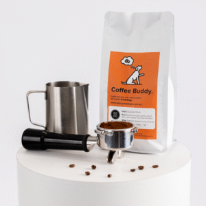 A bag of Soho Coffee beans, a metal milk frothing pitcher, a portafilter filled with ground coffee, and a tamper sit on a white cylindrical surface.