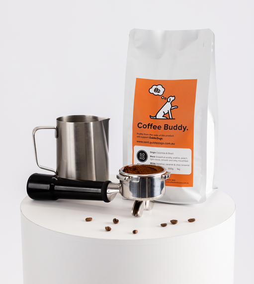 A bag of Soho Coffee beans, a metal milk frothing pitcher, a portafilter filled with ground coffee, and a tamper sit on a white cylindrical surface.