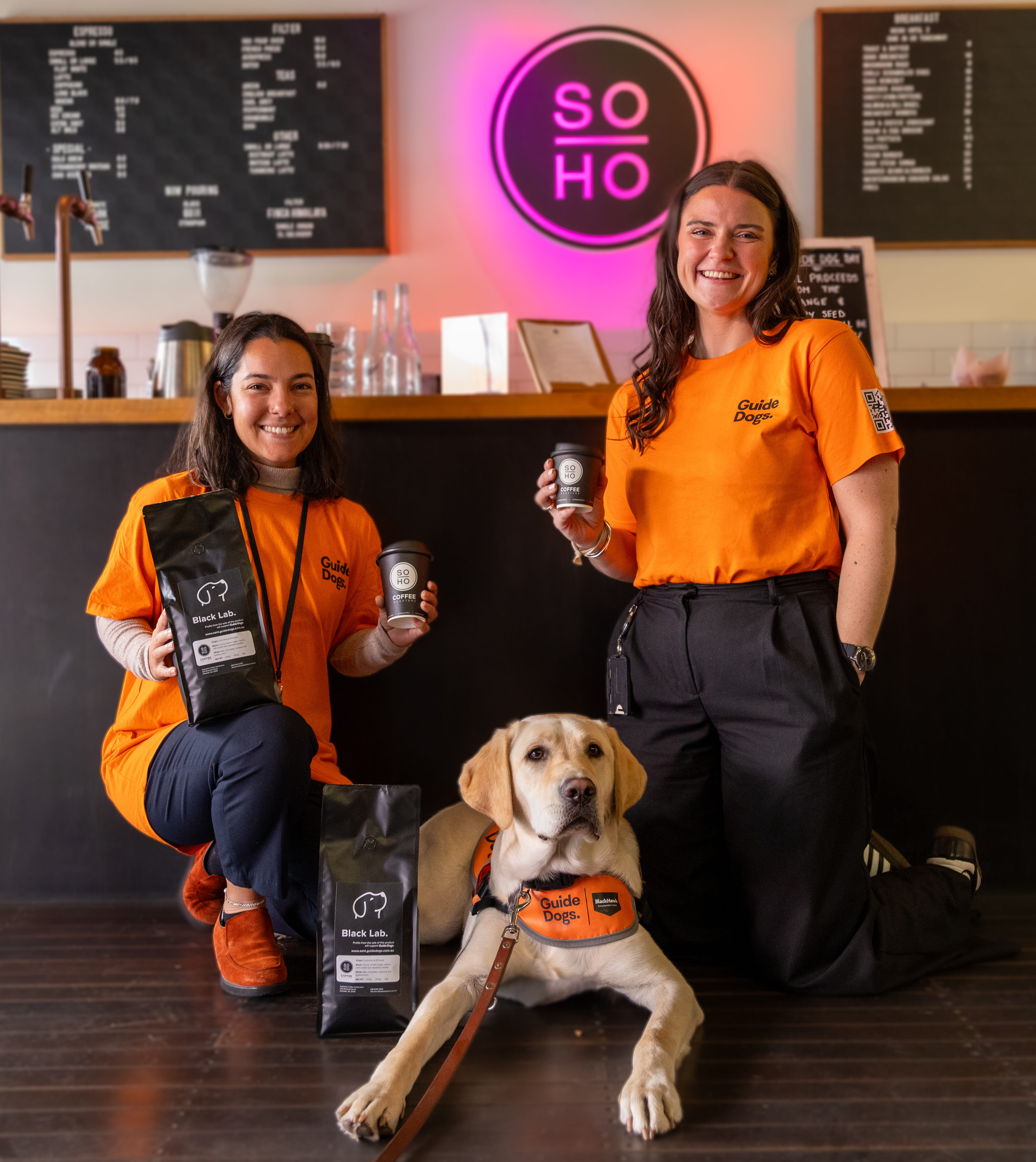 Two women in orange shirts kneel by a guide dog in front of a café counter, holding Soho Coffee and Hot Chocolate, with a neon SOHO sign and menu boards visible in the background.