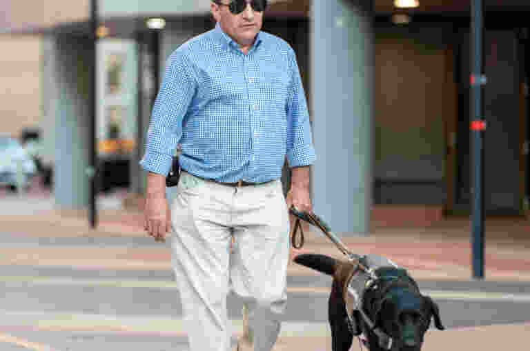 Client, David, walks on the pavement of the Adelaide CBD, guided by black Guide Dog Grant on a harness.