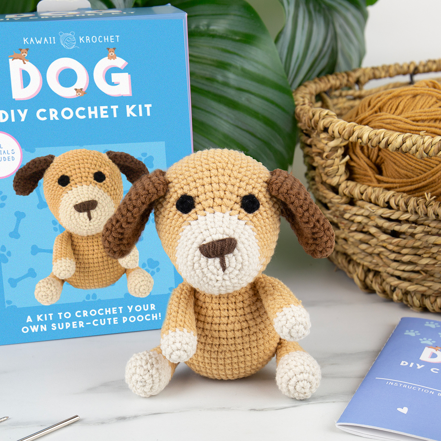 A crocheted dog plush sits in front of a Crochet Kit box, with yarn and crochet tools visible in the background.