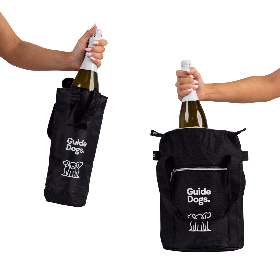 Two hands each hold a black Guide Dogs branded Wine Cooler Bag containing a bottle, one being tote-style and the other a zippered pouch.