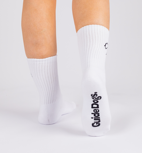 A person wearing Crew Socks - White with Guide Dogs printed in black on the sole, standing on a white background.