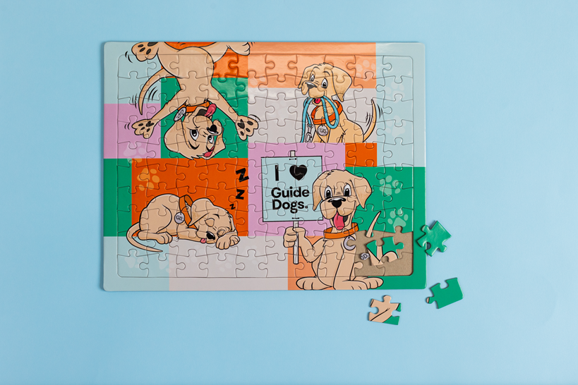 A completed Jigsaw Puzzle featuring a cartoon dog and “I ❤️ Guide Dogs” in the center on a light blue background, with three pieces lying beside it.