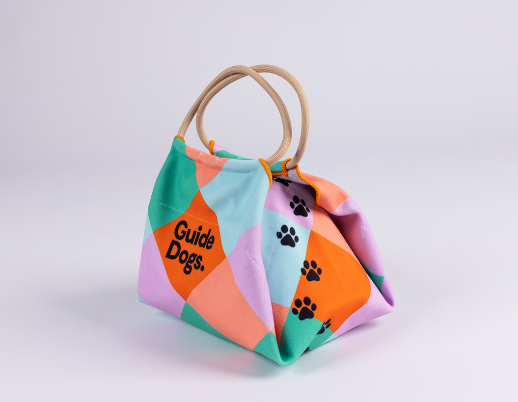 A pastel patchwork Lunch Bag with a handle, black paw prints, and “Guide Dogs” text.