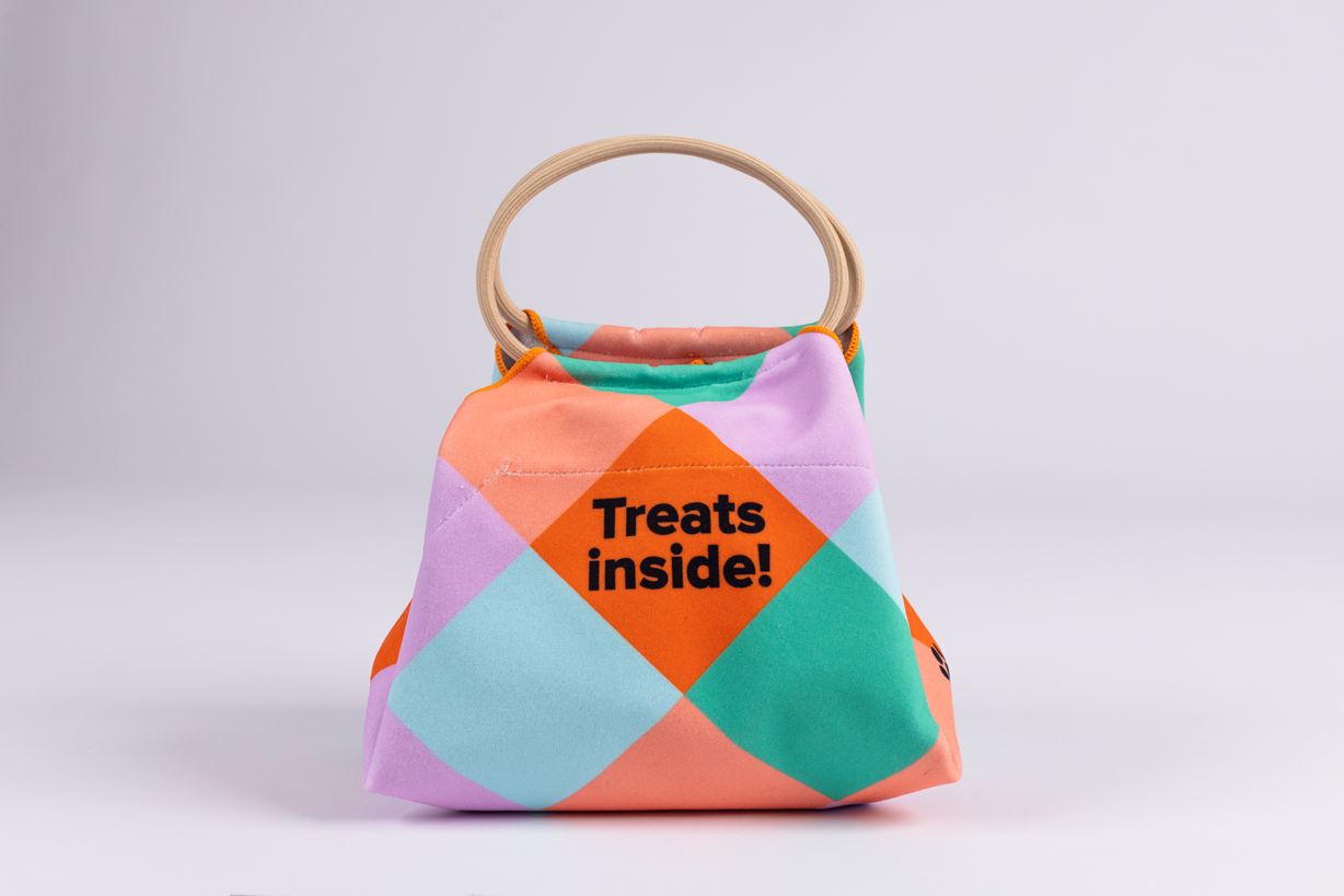 The Lunch Bag is a small, colorful fabric bag with a handle and geometric pattern. The front displays the text Treats inside!.