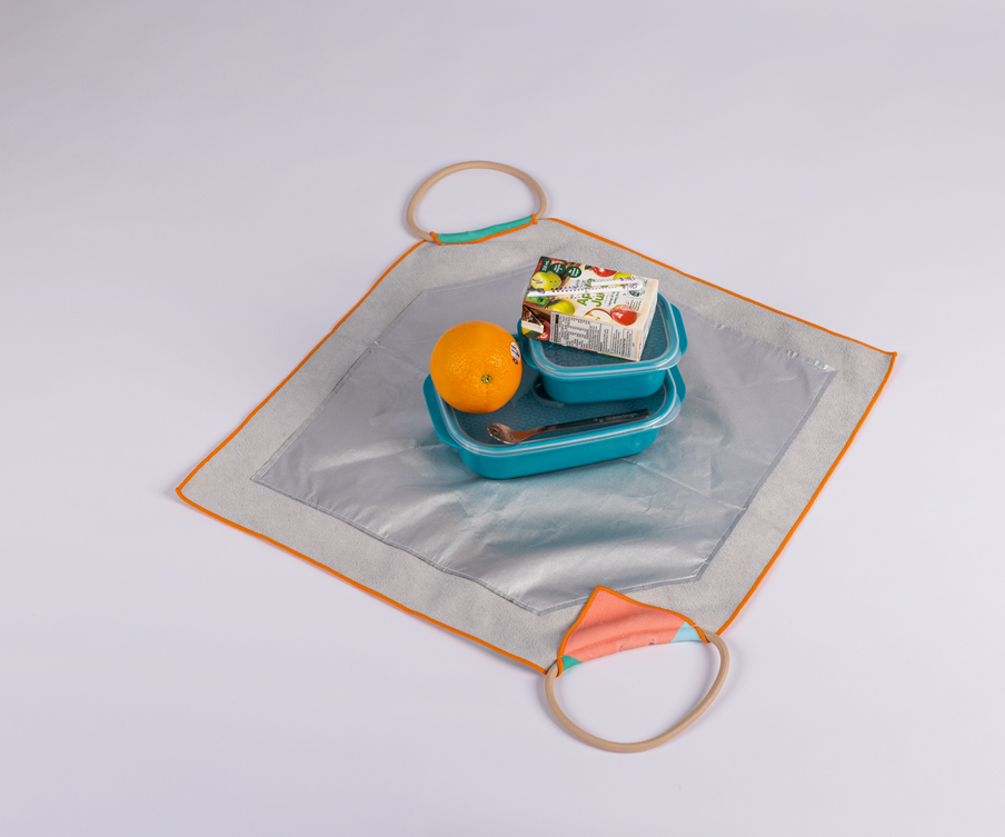 A silver square lunch bag with orange trim and two corner handles holds an orange, a packaged snack, and a blue Lunch Bag with a fork.