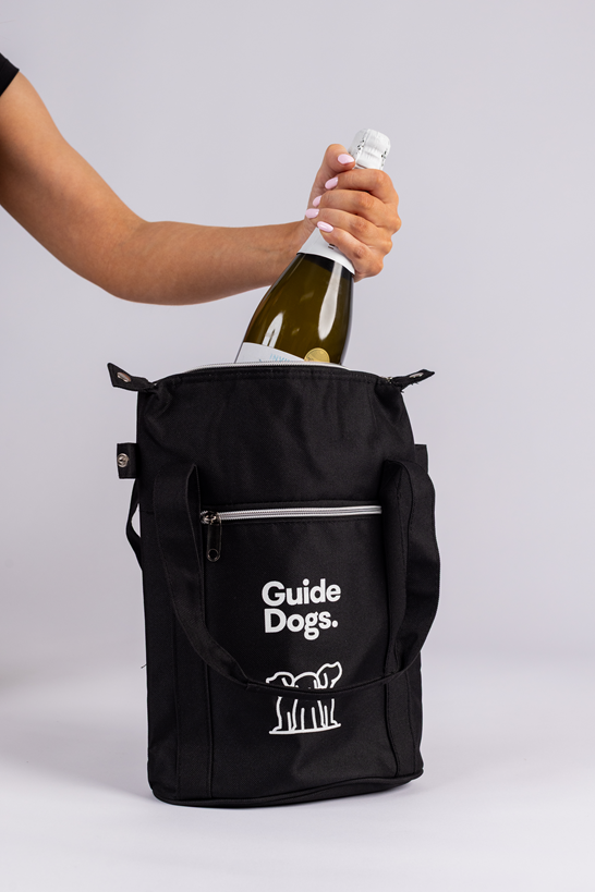 A person puts a wine bottle into a Wine Cooler Bag, which is black and labeled Guide Dogs.