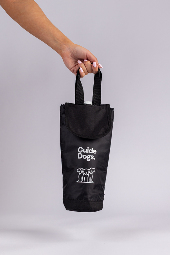 A hand holds a black fabric Wine Cooler Bag with Guide Dogs and a guide dog logo printed on it, set against a plain light grey background.