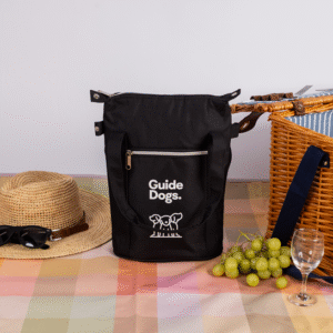 A black Wine Cooler Bag sits on a plaid tablecloth next to a straw hat with sunglasses, a wicker picnic basket, green grapes, and two empty wine glasses.