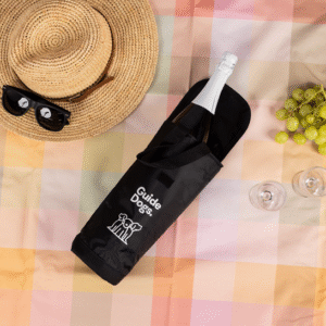 A picnic setup features a straw hat, black sunglasses, green grapes, two empty wine glasses, and a bottle in a black Wine Cooler Bag on a pastel checkered blanket.