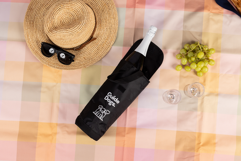A picnic setup features a straw hat, black sunglasses, green grapes, two empty wine glasses, and a bottle in a black Wine Cooler Bag on a pastel checkered blanket.
