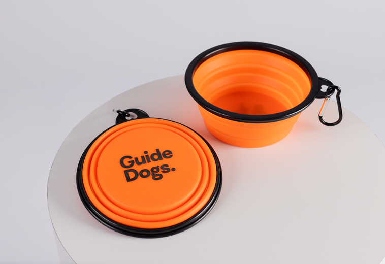 Two orange collapsible bowls with black rims and clips from the Puppy Love Pack, one folded and labeled Guide Dogs, are displayed on a white surface.