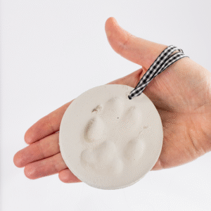 A hand holds the Unboxed but Perfect - Clay Paw Print Kit's round white ornament featuring a paw print impression and a black-and-white checkered ribbon.