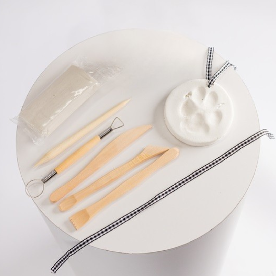 The Unboxed but Perfect - Clay Paw Print Kit includes sculpting tools, a clay packet, and a round clay piece with a paw print, all displayed on a white table with a black-and-white ribbon.