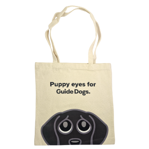 The Tote Bag - Puppy Eyes is a beige bag featuring "Puppy eyes for Guide Dogs." above a cartoon of a dog's eyes and forehead.