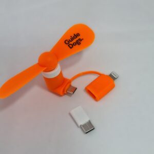 An orange Phone Fan with "Guide Dogs" branding, compatible with Lightning, USB-C, and micro USB connectors, is displayed on a light surface.