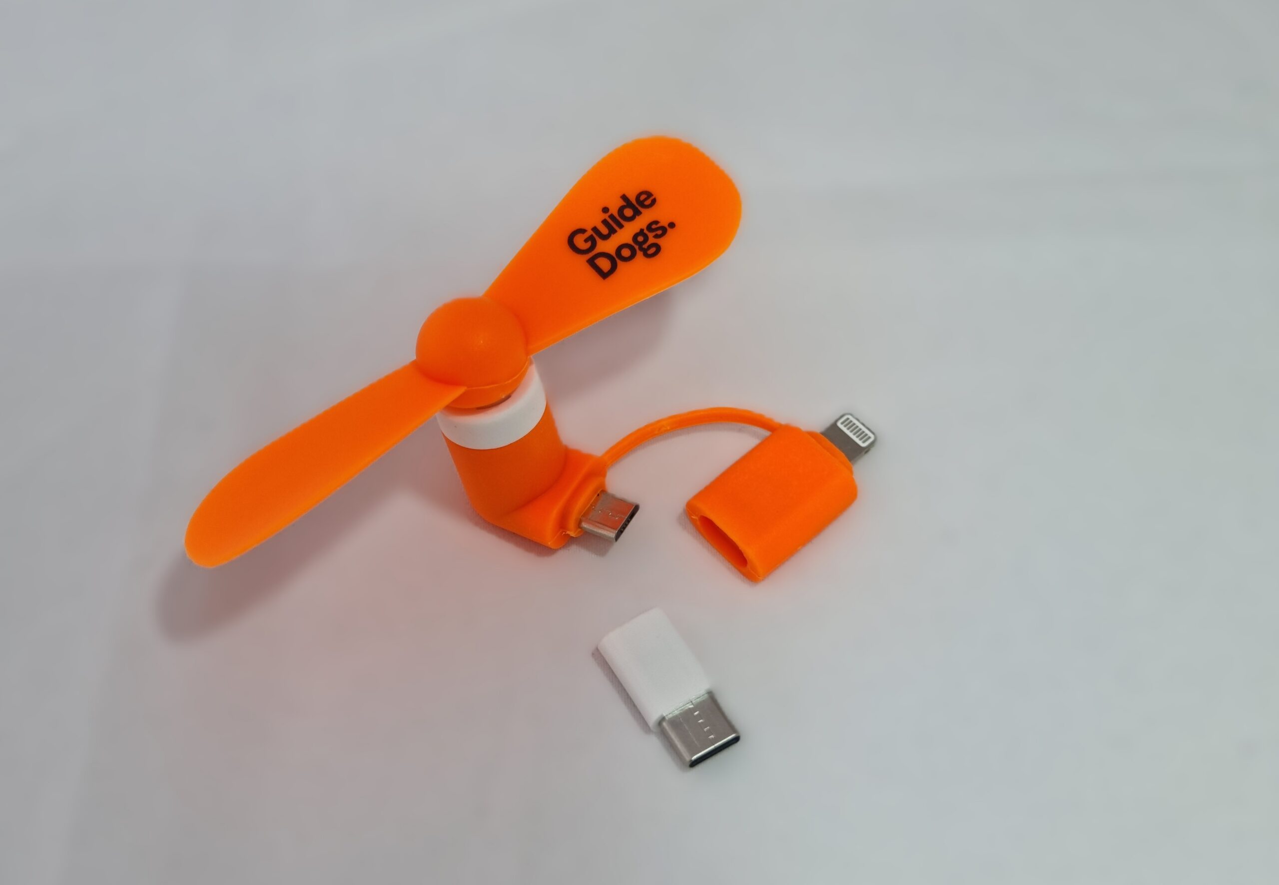 An orange Phone Fan with "Guide Dogs" branding, compatible with Lightning, USB-C, and micro USB connectors, is displayed on a light surface.