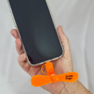 A hand holds a smartphone with a Phone Fan—a small orange fan labeled Guide Dogs—attached to the charging port.