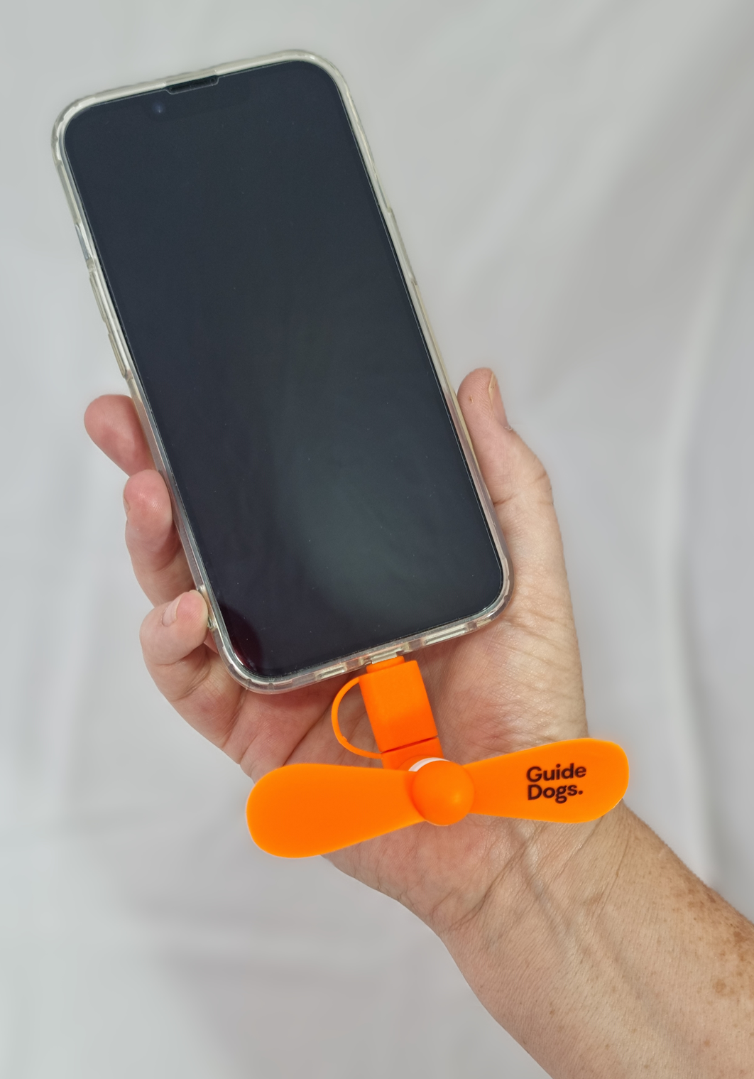 A hand holds a smartphone with a Phone Fan—a small orange fan labeled Guide Dogs—attached to the charging port.