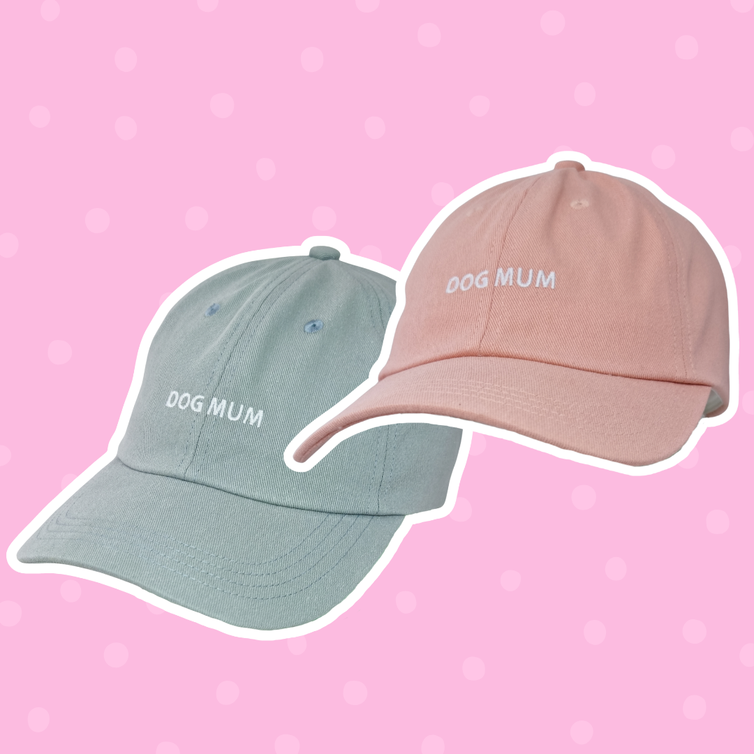 Two Caps, one light blue and one light pink, both with "DOG MUM" embroidered in white, displayed on a pink polka dot background.