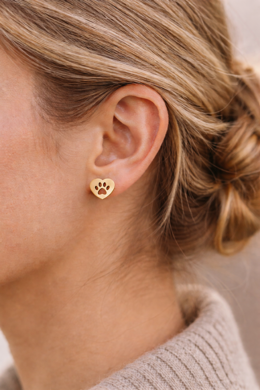 Close-up of a person with light brown hair in a low bun, wearing Earrings - Heart: gold earrings shaped like hearts featuring a cut-out paw print design.