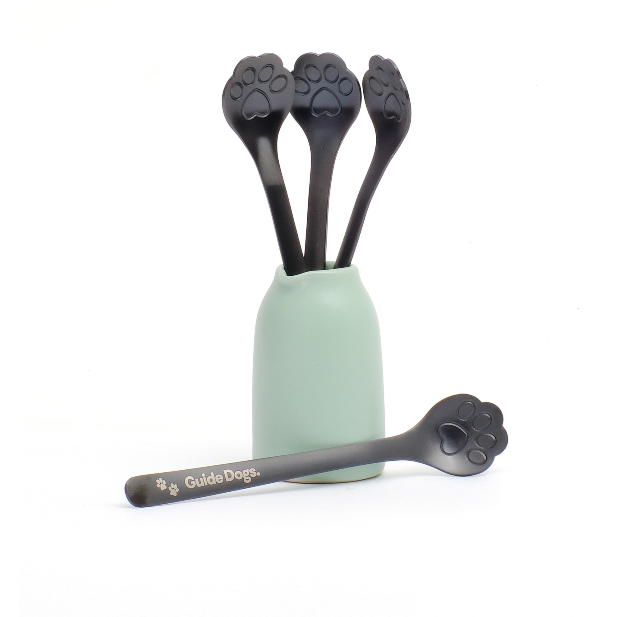 A mint green cup holds four Paw Spoon - Black, each with paw-shaped ends; one matching spoon lies in front, labeled Guide Dogs with a paw print.